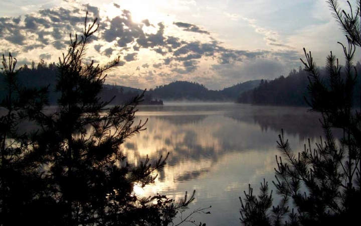 Gentle light is reflected on still water, surrounded by hills and trees.